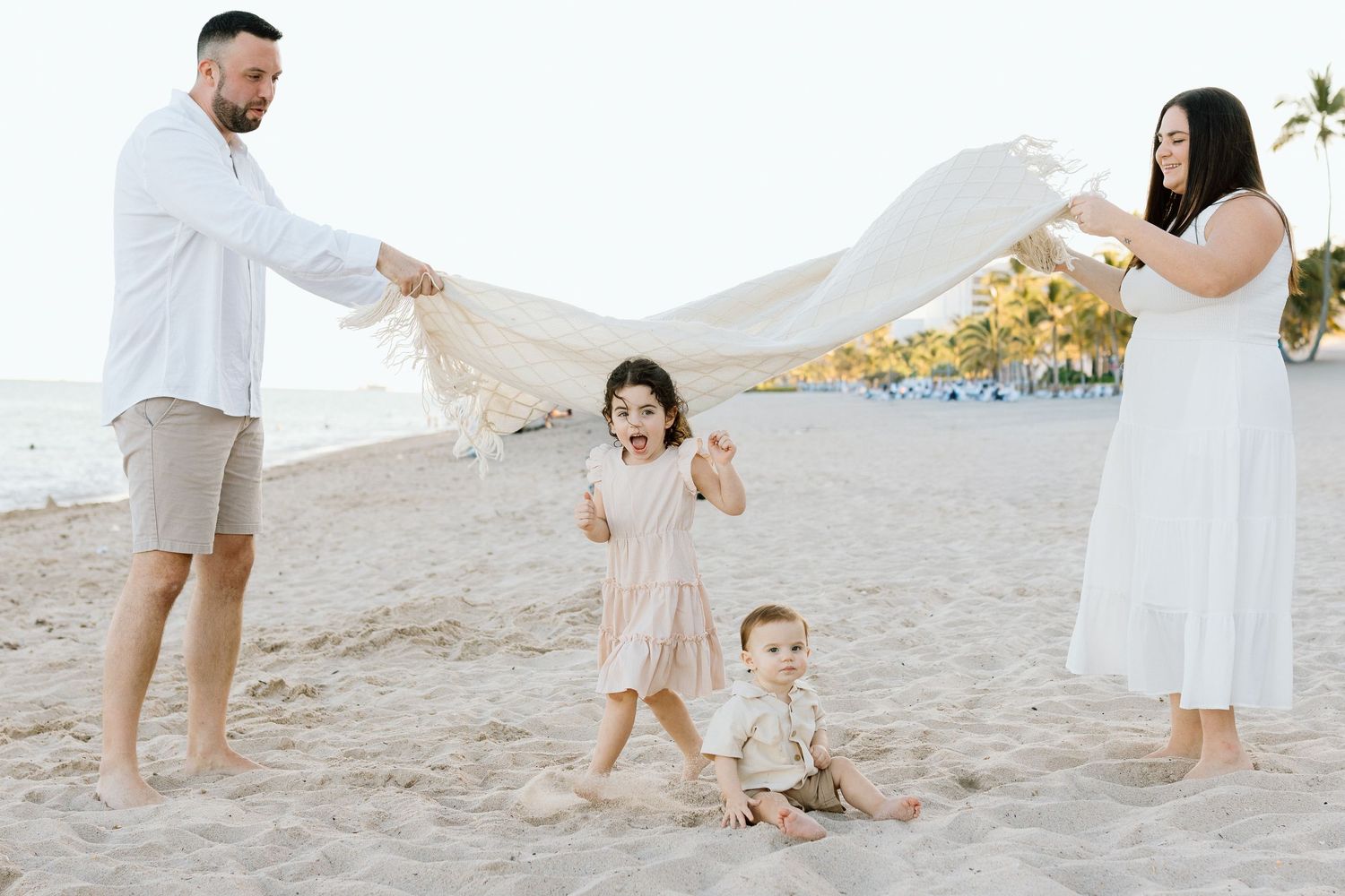 Family of four playing on a sandy beach, parents waving a white blanket over two young children in soft neutral outfits.