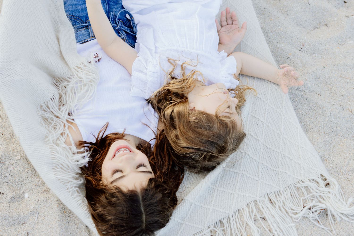 Two smiling women lying on a blanket on the beach, enjoying a sunny day outdoors together.