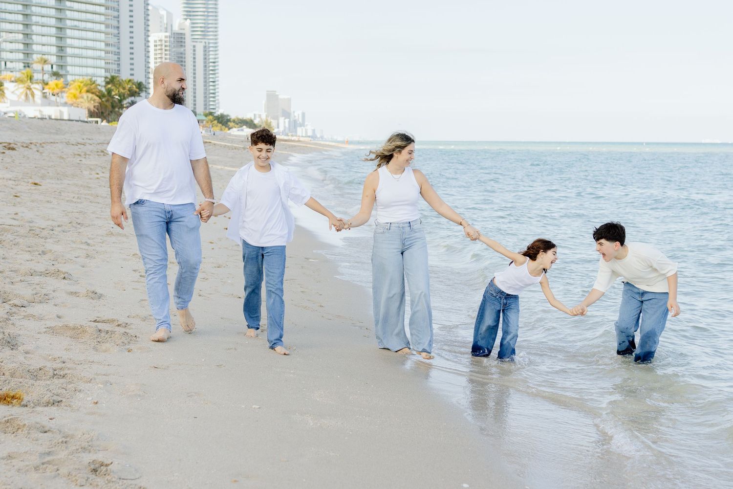 Family of five holding hands walking along a sunny beach with city skyline in the background.