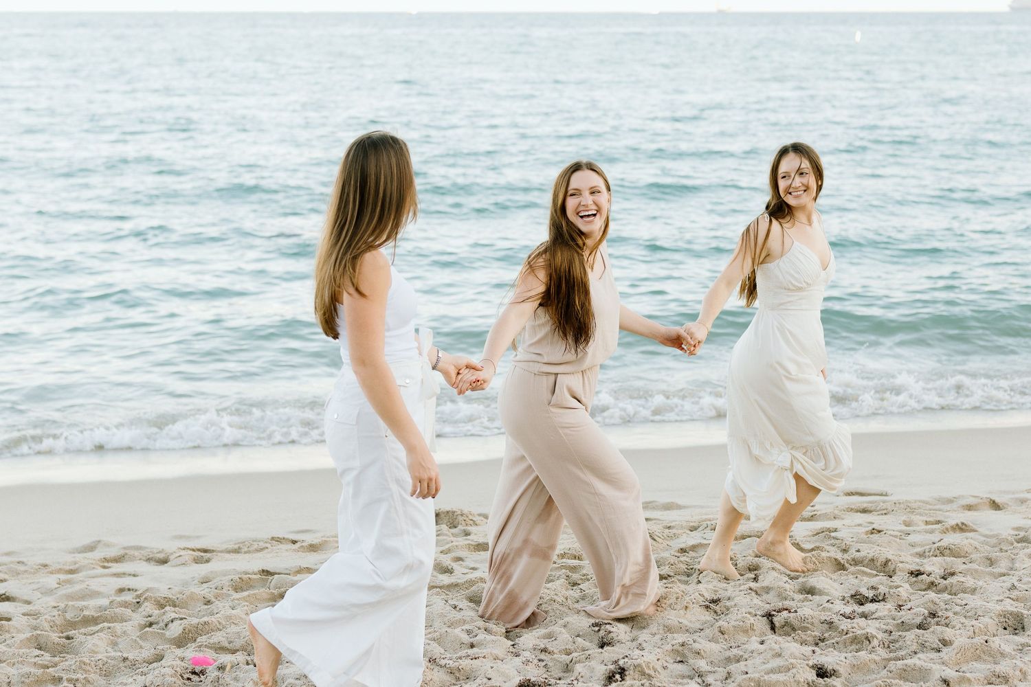 Three women in flowy dresses laugh and hold hands while dancing on a sandy beach near the ocean.