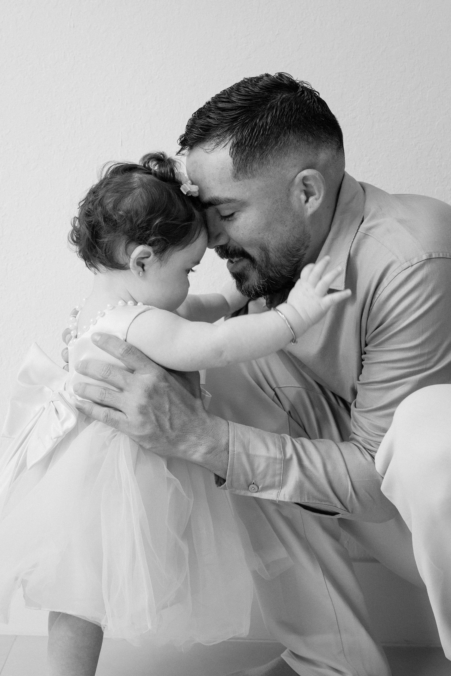 Black and white photo of a father and toddler daughter in a white dress sharing a tender forehead-to-forehead moment.