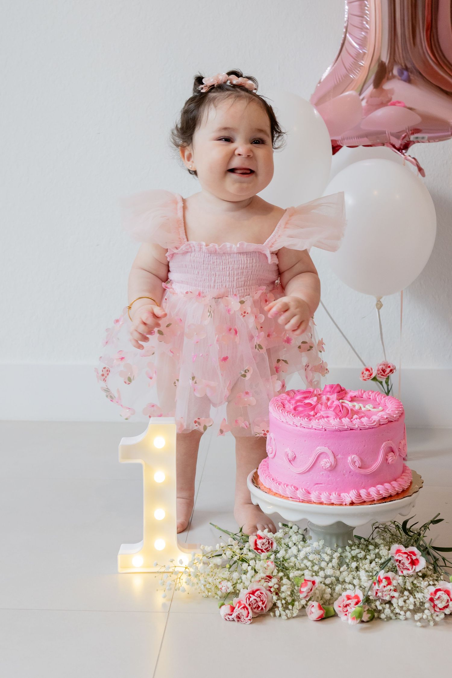 Baby girl in pink dress celebrates 1st birthday with pink cake, balloons, and floral decorations.