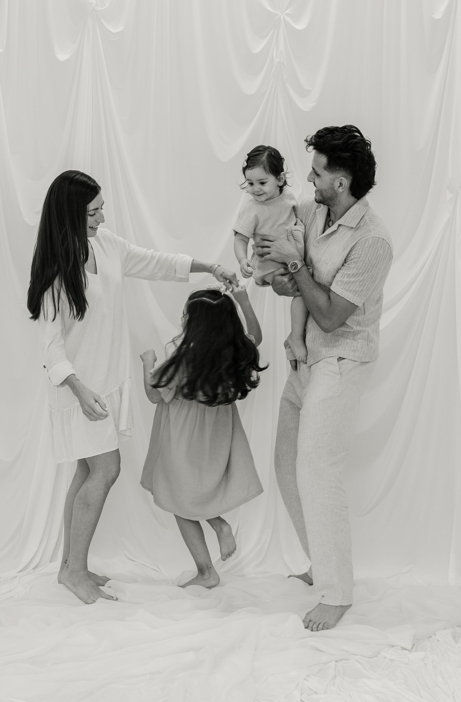 Black and white family photo of parents and two young children playing together in a bright studio setting.
