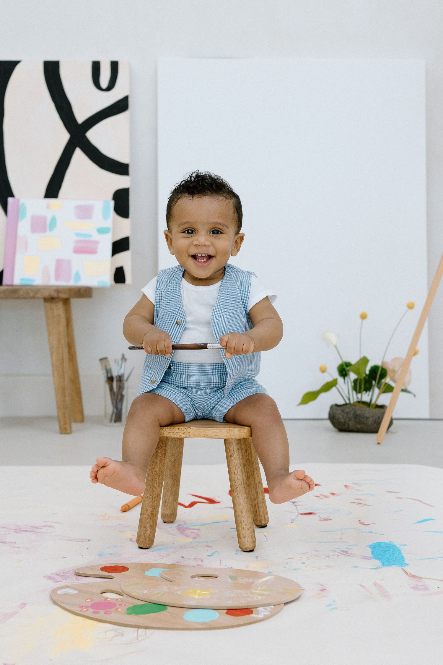 Smiling baby in blue outfit sitting on wooden stool surrounded by colorful paint splatters in an art studio.