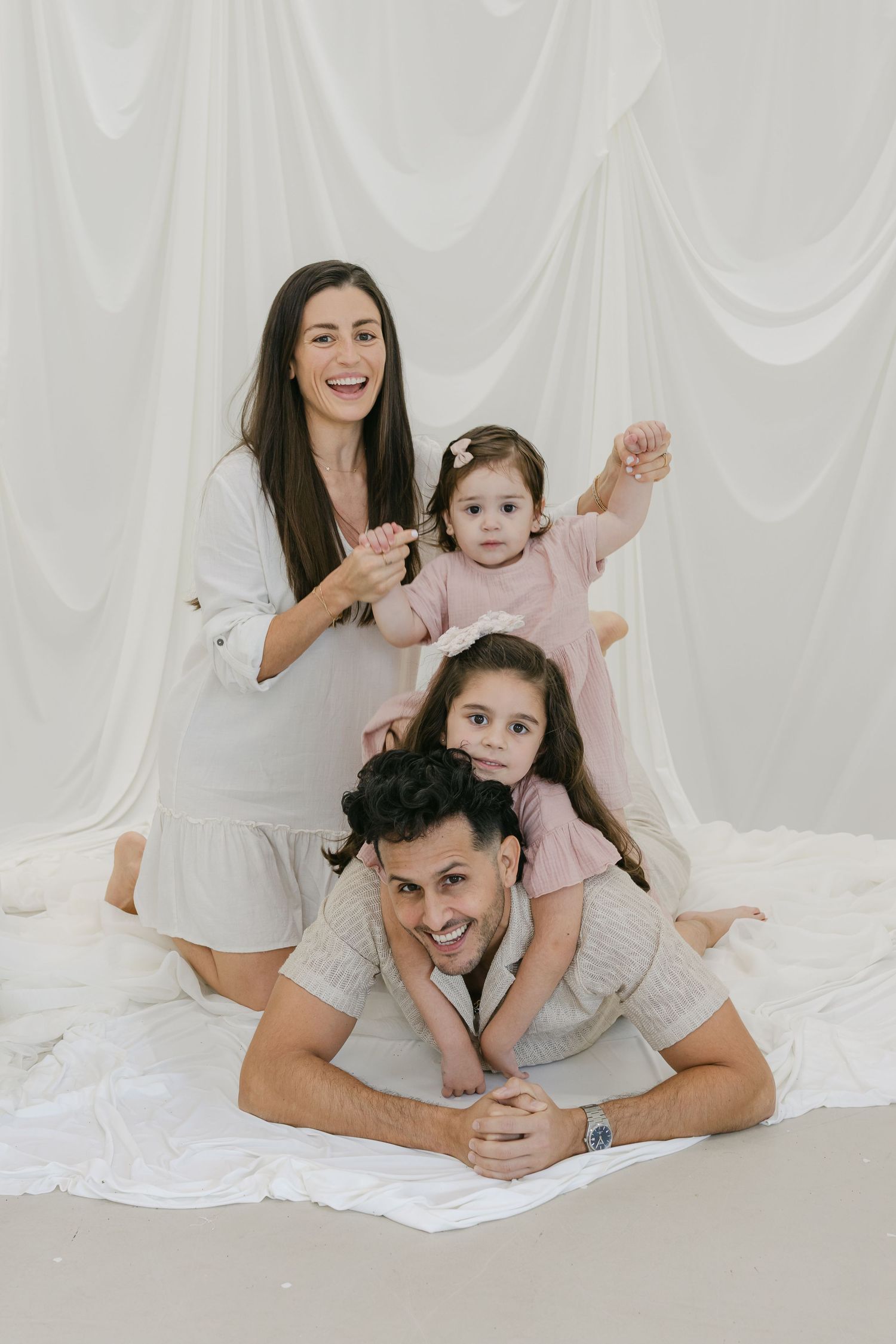 Happy family of four posing in a bright white studio, with mom, dad, and two young daughters in soft pastel outfits.