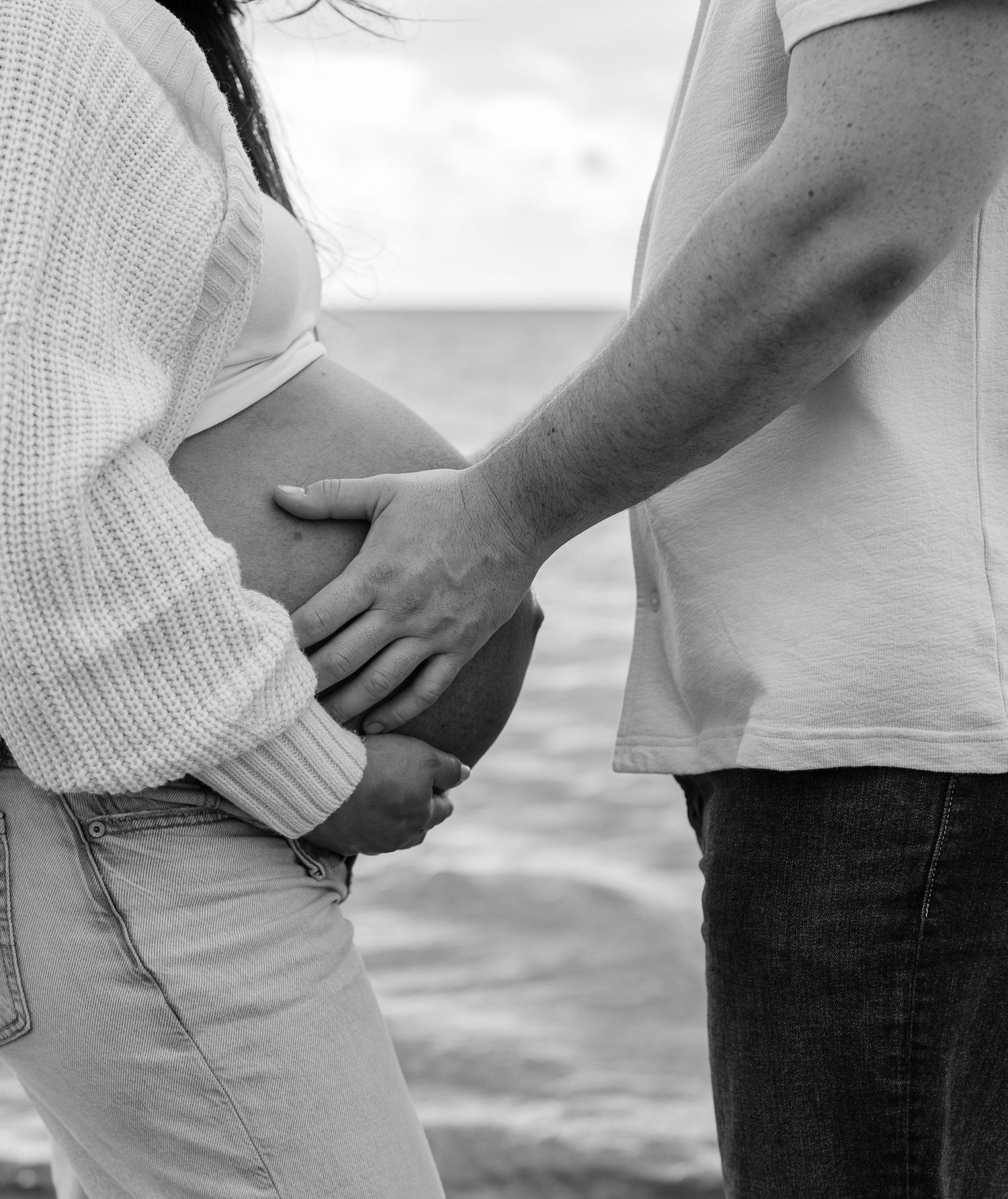 Black and white photo of a pregnant woman and partner holding her baby bump at the beach.