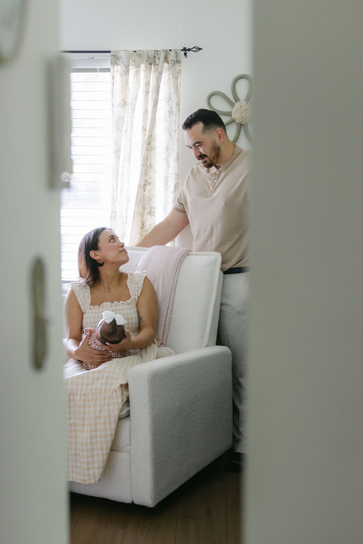 A couple shares a tender moment in a nursery, with the woman seated holding a newborn baby while the man stands beside her.