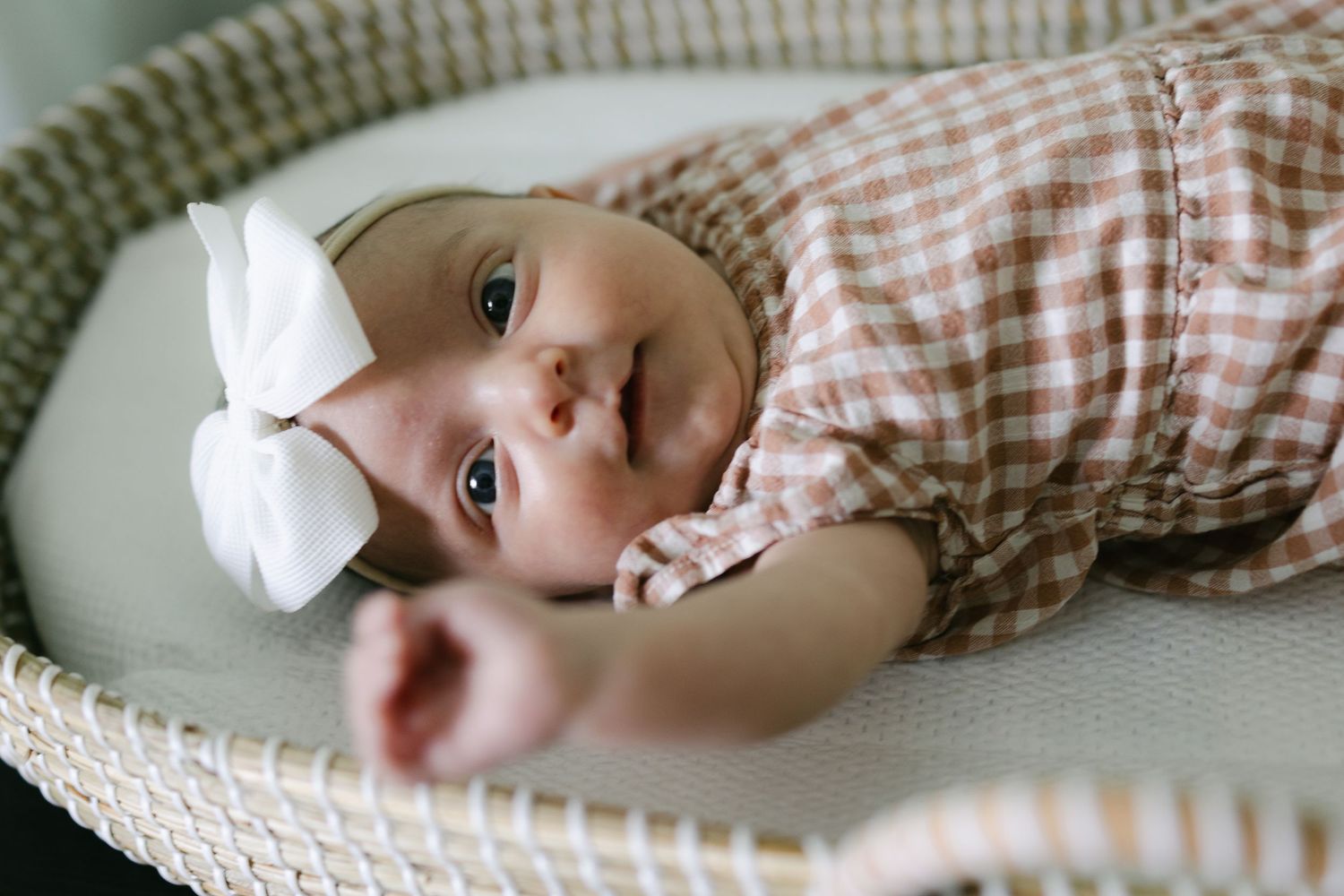 Adorable newborn baby girl in a pink checkered outfit and white bow headband lying in a bassinet.