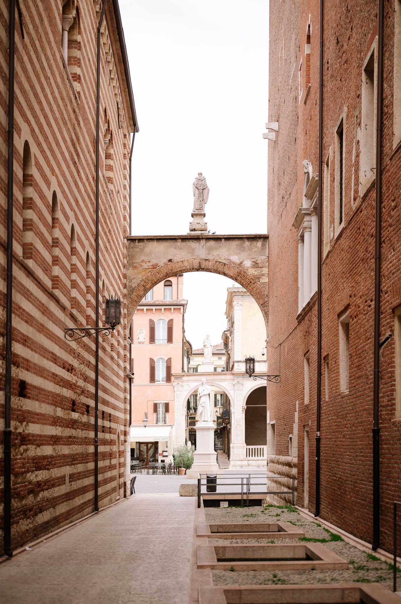 Narrow cobblestone alley between historic brick buildings leading to an arched gateway with a statue in an Italian city.