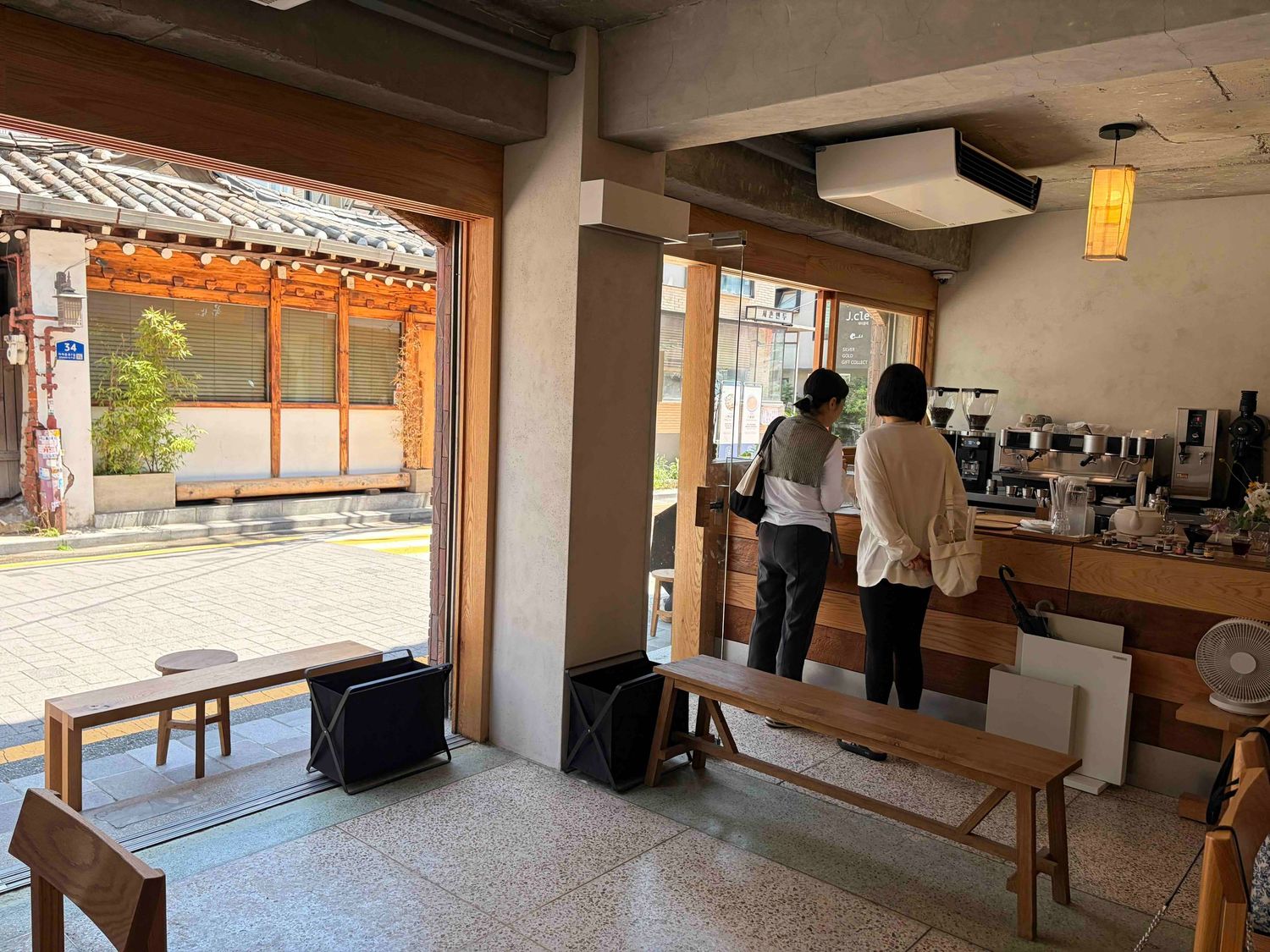 Two people standing at a wooden counter in a minimalist Japanese cafe with open storefront and courtyard view.