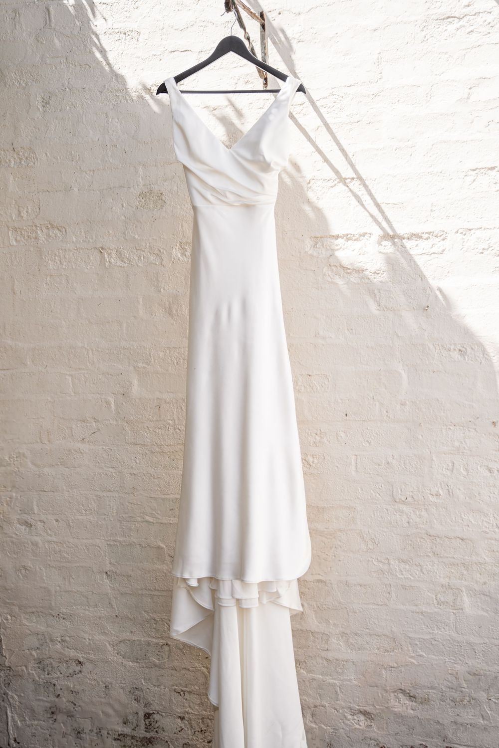 Elegant white off-shoulder wedding dress hanging on a hanger against a white brick wall.