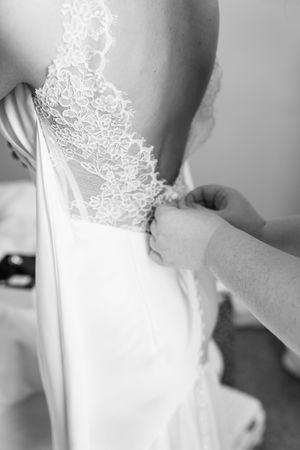 Black and white photo of hands fastening lace back wedding dress buttons on bride.