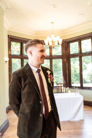 Groom in dark velvet suit with burgundy tie and boutonniere smiling in elegant wedding venue.