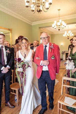 Bride in white gown walks down aisle with man in red blazer at elegant indoor wedding ceremony.
