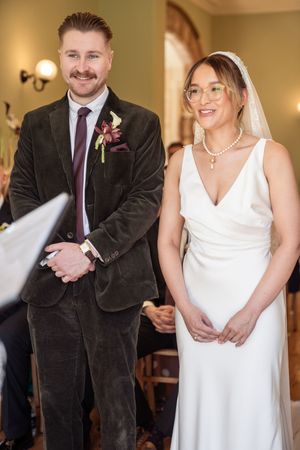 Smiling groom in velvet suit stands beside bride in white gown and veil during wedding ceremony.