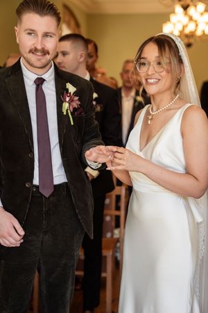Smiling bride and groom exchange rings during wedding ceremony, bride wearing white gown, veil, pearls, and glasses.