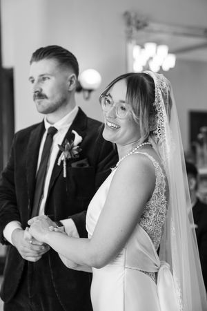 Smiling bride in pearl necklace and veil holds hands with groom in dark suit at wedding ceremony.