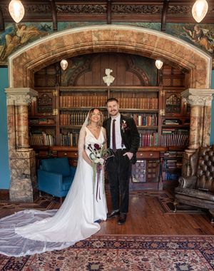 Bride and groom pose in an ornate library with arched bookcase and decorative murals on their wedding day.