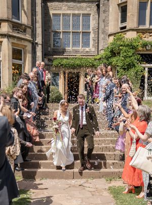 Bride and groom exit a stone manor house as guests throw white confetti on their wedding day.