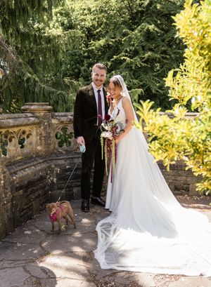 Bride and groom posing with small dog on leash in lush garden wedding venue.