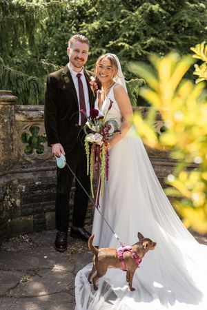 Smiling bride and groom pose with small chihuahua on leash in garden, bride holding colorful bouquet in white gown.