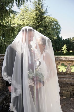 Bride and groom share a romantic moment hidden beneath a lace wedding veil outdoors.