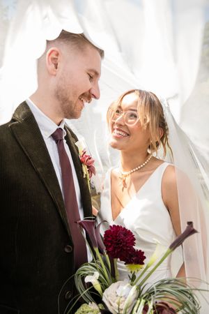 Smiling bride in white dress and veil with groom in dark suit sharing a romantic moment under wedding veil.