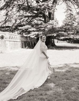 Bride in elegant white gown and long veil twirling in garden, black and white wedding photo.