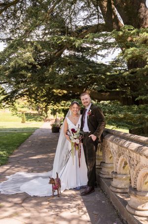Bride and groom pose with small dog on stone pathway surrounded by lush trees at elegant outdoor wedding venue.
