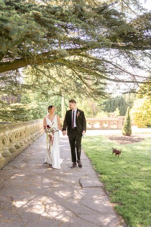 Bride and groom walk hand in hand with small dog along scenic tree-lined pathway at outdoor wedding venue.