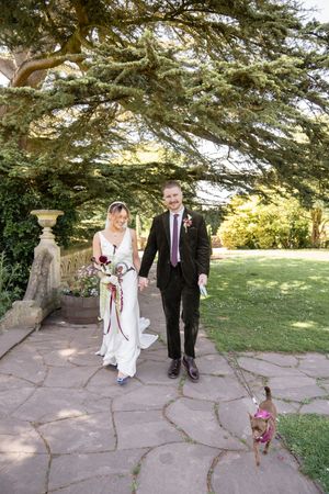Smiling bride and groom walking hand in hand on stone path with small dog at wedding venue.