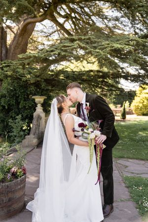 Bride and groom share romantic kiss in lush garden, bride holding burgundy wedding bouquet.