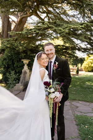 Smiling bride and groom embracing outdoors with flowing veil and cascading bouquet.