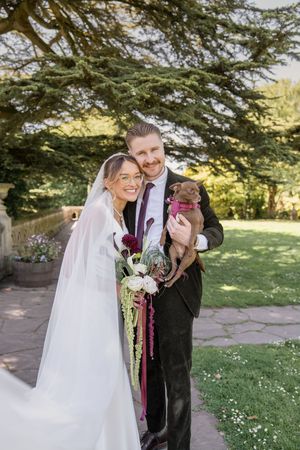 Happy bride and groom posing with small dog in pink outfit at outdoor garden wedding ceremony.