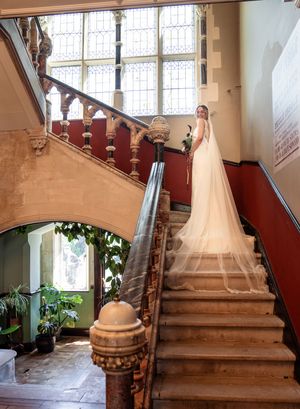 Elegant bride in white gown with long cathedral veil poses on grand ornate staircase in historic venue.