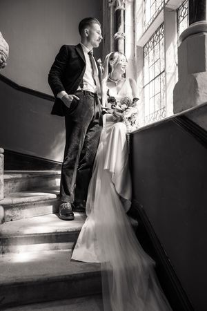 Elegant bride and groom standing on grand staircase by stained glass window, black and white photo.