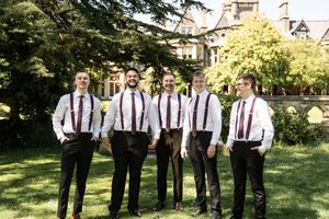 Six groomsmen in white shirts with burgundy suspenders posing on lawn outside manor.
