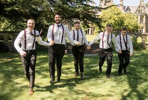 Five groomsmen in black trousers and suspenders walk confidently across green lawn outside a grand stone manor house.