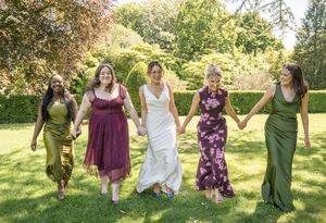 Bride and four bridesmaids in colorful gowns walk hand-in-hand across a sunny green lawn.
