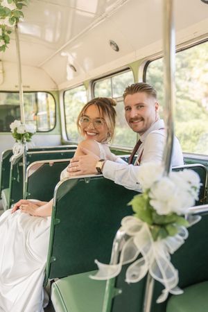 Newlywed couple sits smiling on a vintage green bus decorated with white wedding flowers.