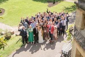 Large wedding party group celebrating outdoors, cheering and raising hands in sunny garden setting.