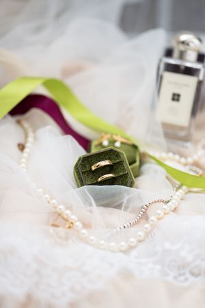 Gold wedding bands in green velvet ring box with pearl necklace and Jo Malone perfume on lace veil.