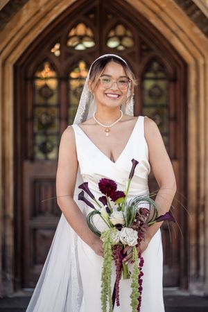 Bride in white gown and veil holds dark floral bouquet while posing before ornate wooden doors.
