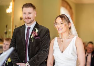 Bride in white gown with glasses and groom in velvet suit smiling during ceremony.