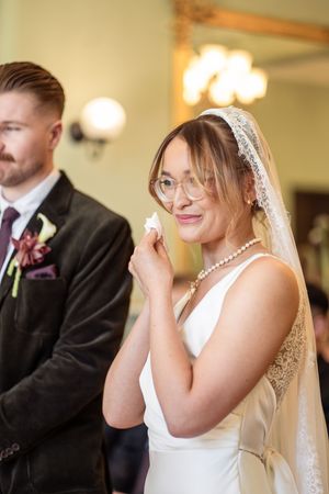 Emotional bride in lace veil and glasses dabs tears during indoor wedding ceremony.