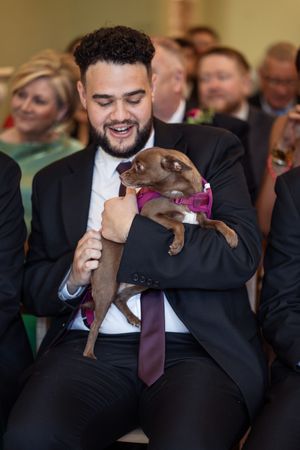 Smiling groomsman holds a small brown Chihuahua in a pink bow tie at a wedding ceremony.