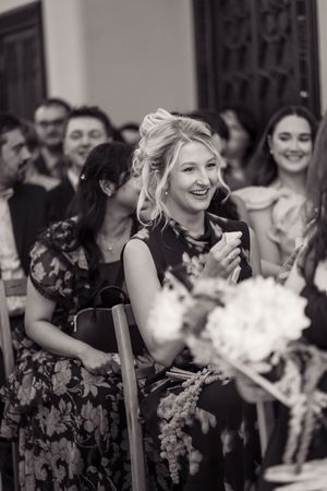Smiling wedding guest in floral dress applauds during ceremony surrounded by other guests and floral arrangements.