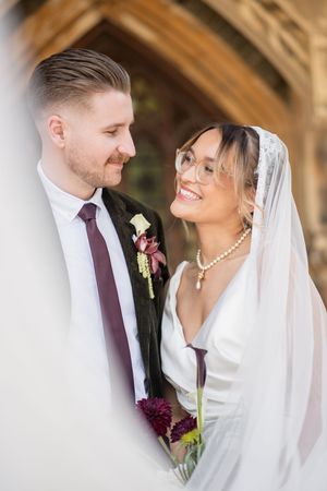 Bride in white gown and veil smiling at groom in dark suit with burgundy tie outside church.