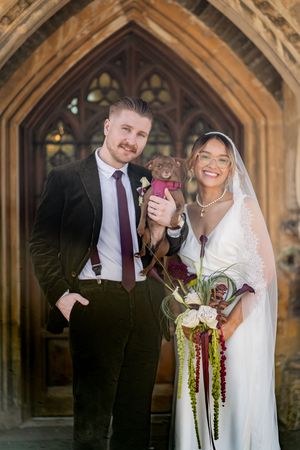 Bride and groom holding small brown dog posing before ornate Gothic church doors on wedding day.