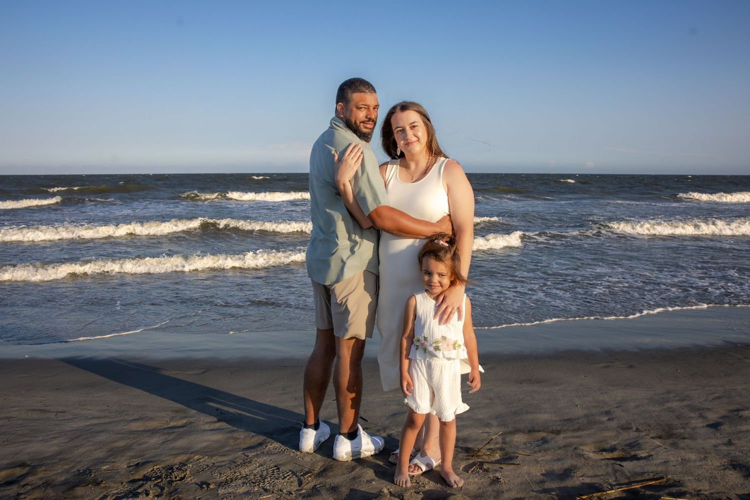 Family of three smiling together on a sandy beach with ocean waves in the background on a sunny day.